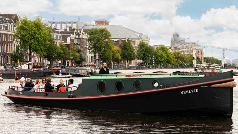 Un barco turístico con pasajeros navega bajo un puente de arco de piedra en los canales de Ámsterdam, mientras los peatones caminan por el puente.